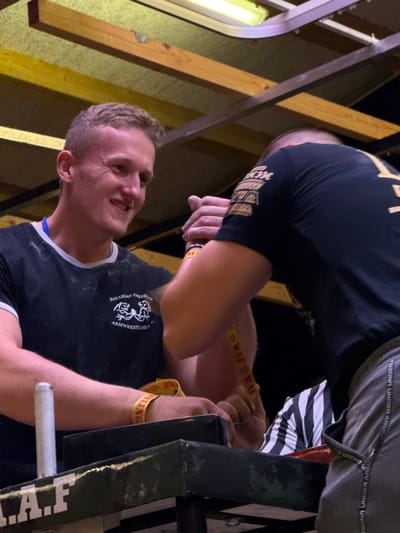 This image depicts an indoor armwrestling match in Reisenberg, Austria. Two male competitors are engaged at a dark-colored armwrestling table, secured by a yellow strap marked "WAF" around their clasped right hands.
The participant on the left, a light-skinned male with short light-brown hair, is smiling. He wears a black t-shirt with white trim and a "BAVARIAN GRIZZLYS ARMWRWESTLING" logo, a blue lanyard, and a yellow wristband. White powder is visible on his left arm and shirt. His left hand is positioned near a vertical metal peg on the table.
The participant on the right is partially visible from the back, wearing a black t-shirt with gold/tan graphics on the right sleeve and grey athletic shorts with "UNDER ARMOUR" text. A referee in a black and white striped shirt is partially visible between the competitors, beneath their joined arms. The table features black padded elbow rests, and "I.A.F" is partially visible in white text on its front edge. Overhead, fluorescent tube lights illuminate a utilitarian ceiling structure with wooden beams. The background is dark.
