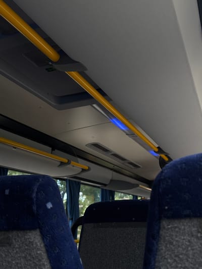 The image shows the interior of a bus or train. The primary subjects are the seats and grab bars. The seats appear to be upholstered in dark blue and grey fabric. Yellow grab bars run horizontally along the wall of the vehicle, indicating it is designed for standing passengers. The scene is brightly lit, with a blue light strip visible above the grab bar, suggesting interior lighting. The windows of the vehicle are visible, showing a blurry, green outdoor scene with trees, suggesting a daytime setting in a possibly rural or suburban area. There are no people visible in the image. The overall impression is that of an empty public transport vehicle in motion. No text is visible in the image. The location context provided is Olaine, Latvia.