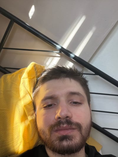 A close-up, angled shot features a man lying down on a yellow pillow with a dark, possibly iron, bed frame visible in the top left. The man has short brown hair, a beard, and appears to be looking at the camera. The background is a white wall with sunlight streaming through blinds or a window, creating light and shadow patterns. The location, Pentre, United Kingdom is unknown and could be a domestic setting.