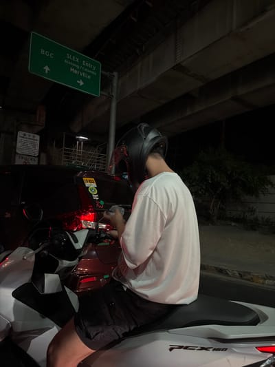 A man wearing a black helmet, white shirt, and black shorts is straddling a parked Honda PCX 160 scooter in the city of Pasay, Philippines. He is bending over the scooter's instrument panel. A green street sign indicates "SLEX Entry Alabang/Calamba Merville," with arrows pointing to BGC and Merville. The scene is located under an elevated structure, possibly a bridge or highway overpass. A "Trucks" sign is visible in the lower left corner.