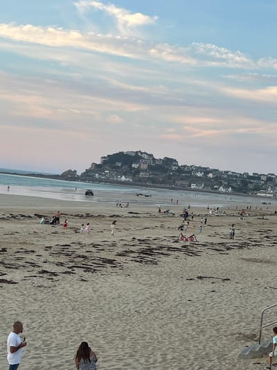 The image depicts a beach scene at Pléneuf-Val-André, France. The sandy beach is populated with numerous people engaged in various activities. In the foreground, a man in a white t-shirt and jeans stands holding an object, while a woman with long dark hair wearing a black and white patterned top stands in front of him. Further back on the beach, several groups of people are visible. Some are sitting on the sand, while others are walking or playing. A tractor is present on the wet sand near the waterline. In the background, a town with white buildings is nestled on a hill overlooking the sea. The sky above is filled with soft clouds, tinged with pink and yellow hues, suggesting it might be late afternoon or early evening. The overall scene conveys a relaxed atmosphere of a day at the beach.