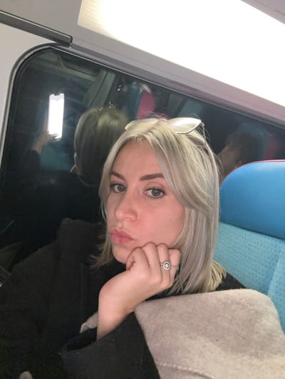 A young woman with blonde hair and light eyes poses for a selfie. She's wearing a black coat and a large ring. Her hand is resting on her cheek. The background appears to be a train, visible through the window, with reflections showing the woman and a glowing screen. The train seat is blue and grey. The image is rotated slightly.