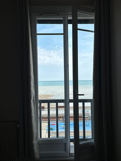 This image captures a view through an open French door, looking out towards the sea from a room in Saint-Valery-en-Caux, France. The foreground is dominated by dark gray curtains on either side, framing the doorway. To the left, partially visible, is a radiator. The open door reveals a second layer of glass panels with a black railing in front of them. Beyond the railing, a vibrant blue surface, likely a playground, is visible with red and green play structures. Further out, the ocean stretches to the horizon under a pale blue sky with scattered clouds. The shoreline is rocky, with a narrow strip of pebbles meeting the water. The overall impression is one of a tranquil seaside scene viewed from an interior space.
