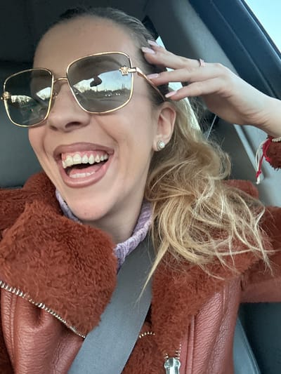 A blonde woman in a car wearing a brown fur-lined jacket, a seatbelt, and large square sunglasses is seen smiling at the camera. She is wearing a ring on her left hand, which is raised to the top of her head. It is not possible to determine her location in the city of Basildon, United Kingdom, based on the image.