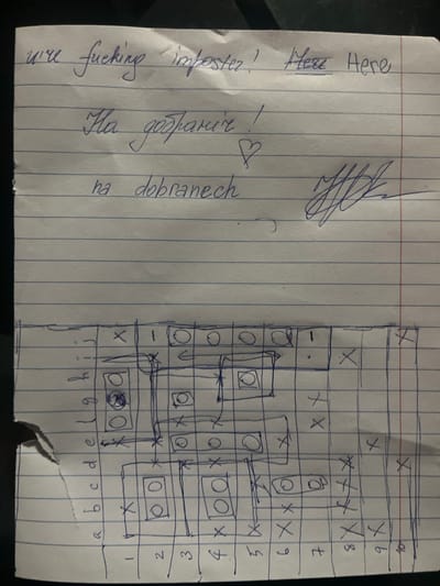 A creased, white, blue-lined notebook paper, approximately 8.5 x 11 inches, is depicted with handwritten text and a drawn grid in blue ink. The upper section contains three lines of text: "we're fucking imposter! Here Here" (the first "Here" is underlined), followed by "На добраніч !" (Cyrillic for "Good night!"), a small heart symbol, and then "na dobranech" (a transliteration of "good night"). To the right of the Cyrillic and transliterated text is a complex, illegible signature or scribble. The lower section of the paper features a hand-drawn grid, approximately 10 columns by 10 rows, with columns labeled numerically (1-10) and rows labeled alphabetically (a-j, though some labels are partially obscured by a tear). Within the grid, various cells contain circles (some empty, some with an inner circle), crosses ('X'), and small arrows indicating direction. Several rectangular outlines enclose sets of cells, some containing circles. The paper exhibits a tear on its left edge, affecting the visibility of some row labels. The image does not contain details to identify a specific location within the city of Bakuriani, Georgia.