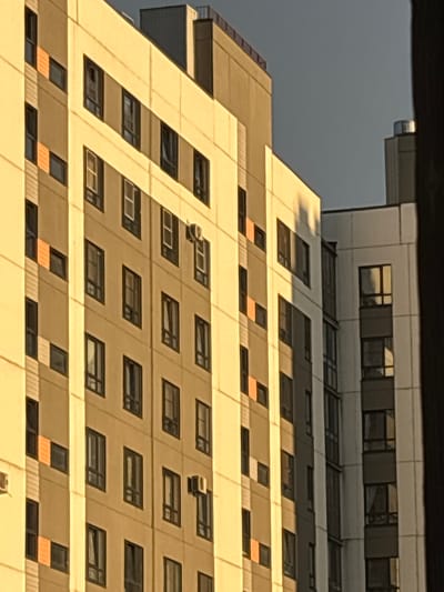 The image shows the side of a multi-story apartment building in Imeni Morozova, Russia. The building features a pattern of light-colored and beige panels, with dark-framed windows and some orange accent panels on the left side. An air conditioning unit is visible on the beige section of the building. The sun is casting a strong yellow light on the facade, creating shadows. To the right of the main building, another section of a lighter-colored building with dark windows is visible, and further to the right, a dark vertical structure obscures part of the view. The sky above is a muted gray. There are no people or animals visible in the image.