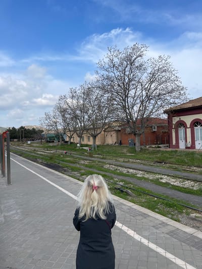 The image depicts a train station in Buñol, Spain, observed from a platform looking down the tracks. In the foreground, a single individual is positioned with their back to the viewer, standing on a gray tiled platform with a white safety line. This individual has long, wavy blonde hair secured with a pink hair clip and is wearing a dark, long-sleeved coat or jacket, with a dark strap visible over their right shoulder.
Parallel to the platform, multiple sets of train tracks are visible, embedded in gravel and partially overgrown with green vegetation. Across the tracks, a series of structures stand. To the immediate right, a building with a red-tiled roof, light-colored walls, and distinctive red trim around two arched windows or doors is present, possibly the main station building. An external air conditioning unit is mounted on its side. Further down the tracks, a line of single-story buildings with light brown walls and dark roofs are visible, interspersed with bare deciduous trees suggesting late winter or early spring. One white plastic chair is discernible near one of these buildings. In the far distance to the left, low hills or mountains are under a partially cloudy blue sky with scattered white clouds.