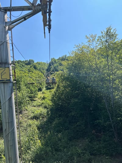 This is a medium-angle, eye-level, outdoor shot of a ski lift in the city of Mizhhiria, Ukraine. In the foreground, on the left, is a section of a large metal ski lift tower with a metal ladder attached. A yellow sign with a black symbol and the word "TATRAPOMA" is affixed to the tower. Above the tower, a steel cable for the ski lift extends upward and to the right. Two people are seated on a ski lift chair, facing away from the camera and traveling up the slope. They are positioned in the middle ground of the image. The background consists of a dense, green, forested hillside under a clear blue sky. The vegetation is thick with trees and shrubs, indicating a summer or late spring season. The ski lift appears to be in operation.