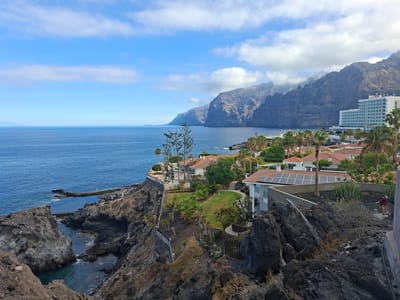 This media file captures a scenic coastal view in Santiago del Teide, Spain. The scene features a dramatic rocky coastline meeting the deep blue ocean under a partly cloudy sky. In the foreground, rough, dark volcanic rocks dominate, with some sparse vegetation growing between them. A man and a woman are visible on a balcony on the right side of the frame, observing the scenery.
Further back, nestled on the clifftop, are several modern buildings with red-tiled roofs, suggesting a residential or resort area. One building prominently displays solar panels on its roof. Lush green gardens and palm trees surround these structures, providing a contrast to the rugged cliffs.
In the distance, majestic, imposing cliffs rise dramatically from the sea, their sheer faces etched with the passage of time. A large white hotel with many floors and blue-tinted windows is situated on the right side of the frame, partially obscured by palm trees and blending with the mountain backdrop. The ocean is calm, reflecting the bright sky, and a small, natural-looking swimming pool is carved into the rocks on the left side of the image, with a few people visible near it.
The time of day appears to be late morning or early afternoon, given the bright sunlight and the angle of the shadows. The weather is pleasant, with a mix of sun and clouds, creating a dynamic sky. The overall impression is one of a beautiful, sun-drenched vacation destination with a striking natural landscape. There is no visible text in the image.