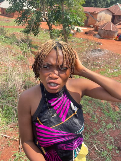 A young Black woman is prominently featured in this vertically oriented photograph, captured outdoors in what appears to be a rural or semi-urban setting in Ogbe, Nigeria. She is likely reclining or lying down, looking directly into the camera with an engaging, somewhat playful expression, her mouth slightly open to reveal her teeth. Her right arm is raised, with her hand near her head, possibly adjusting her hair or posing.
She is wearing a black sleeveless top with a vibrant geometric pattern of fuchsia pink, black, and gold lines and stripes. A delicate golden necklace with an infinity symbol pendant is visible around her neck. Her hair is styled in bleached or light-colored dreadlocks. Part of a yellow garment or item can be seen to her lower right.
The background shows a mix of natural elements and simple architecture under bright daylight. Buildings with rusted, corrugated metal roofs and light-colored walls are discernible behind her, characteristic of local housing. The ground is covered with reddish-brown soil, sparse green grass, and dry vegetation, suggesting a warm and possibly arid or dry season environment. Green foliage and trees are also visible, adding depth to the scene. The image conveys a candid moment within a typical Nigerian landscape.
