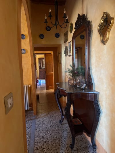The image depicts an interior hallway, likely in Sinalunga, Italy, characterized by antique and decorative elements. The walls are painted in a mottled yellow-orange hue, adorned with decorative blue plates and framed pictures. A chandelier hangs near a large ornate mirror reflecting the hallway's depth and light. An antique wooden console table stands prominently, featuring decorative carvings. In the background, a doorway leads to what appears to be a kitchen or dining area with wooden cabinets and a view of a brightly lit space, perhaps a window. The floor is tiled with a speckled pattern. A radiator is visible along the wall. The scene is still and devoid of people or actions, focusing on the architectural and decorative aspects of the space.