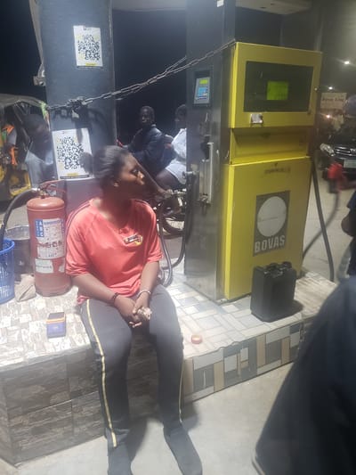 The image depicts a scene at a BOVAS gas station in Alalubosa, Nigeria. A woman in a red shirt and black pants sits on a wheeled chair next to a gas pump with a fire extinguisher nearby. Two men stand near the pump. In the background, a motorcycle is parked, and a black container sits on the gas pump. The gas pump displays a price of 125000 and a volume of 100200. Two QR code stickers are attached to the gas pump. The flooring is tiled.