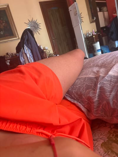 The image shows a close-up view of a person's legs wearing bright orange shorts. The person is lying on a bed with a patterned duvet. In the background, a room is visible with furniture, including a dresser, a wardrobe with clothes, and a refrigerator. A framed painting is on the wall, and a sun-shaped clock is also visible. The overall impression is of a domestic interior. The location in the city of Ardea, Italy, is not discernible from the visual information provided in the image.
