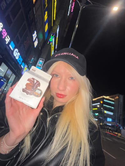 A female subject with long, straight blonde hair and blue eyes is depicted in a close-up, waist-up, nighttime portrait on a city street in Namyangju-si, South Korea. She wears a black baseball cap with "BALENCIAGA" embroidered in pink capital letters, and a black leather jacket with a silver zipper. Her lips are coated with red gloss.

The subject holds a white rectangular carton drink box in her left hand, extended towards the camera. The box features an illustration of a stack of chocolate brownies or bars with chocolate syrup. Korean text on the box includes "설탕 무첨가" (Sugar-free) and "300 mL (150 kcal)". A white wristband and a silver chain bracelet with a small heart charm are visible on her left wrist.

The background is dark, illuminated by numerous vibrant neon and LED signs on multi-story buildings, indicating a commercial district. Prominent among these signs is a partially visible "GS25" convenience store logo. Other Korean signs, including "약국" (pharmacy), display text in various colors like blue, yellow, and pink. A street or sidewalk is visible at the bottom right, with additional city lights in the distance. The overall scene suggests an urban night environment.