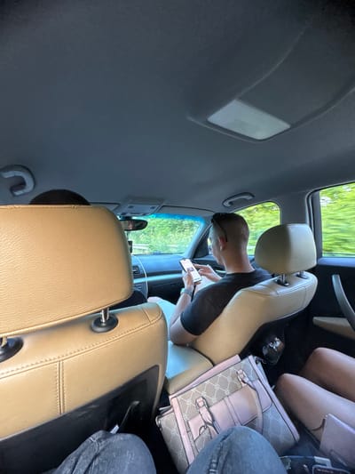 The image captures the interior of a vehicle from a rear-seat passenger's perspective, during transit in Omarchevo, Bulgaria. The car's interior features beige leather seating and a gray headliner.
In the immediate foreground on the left, the upper portion of a beige leather front seat headrest is visible. Behind it, the top of a driver's head, covered in dark hair or a dark cap, is partially discernible.
To the right, a male front passenger is seated, characterized by short hair and sunglasses resting on top of his head. He is wearing a dark t-shirt and is focused on a smartphone, which he holds with both hands. The passenger's seat is also upholstered in beige leather.
The vehicle's ceiling displays a gray headliner with an overhead light fixture, featuring a rectangular, translucent cover, and an adjacent panel of controls. A white grab handle is affixed to the left side of the ceiling. Through the front windshield, a dark-colored dashboard is visible, from which a white cable extends downward. A rearview mirror hangs centrally above the dashboard.
The exterior environment, visible through all windows, is blurred green, indicative of trees and foliage, suggesting the vehicle is in motion along a road.
In the rear passenger area, the legs of at least two individuals are visible. On the left, legs clad in dark jeans are present. On the right, lighter-skinned legs are observed near a light pink, patterned handbag and a smaller pink accessory, both resting on the seat or floor. Natural daylight illuminates the scene.