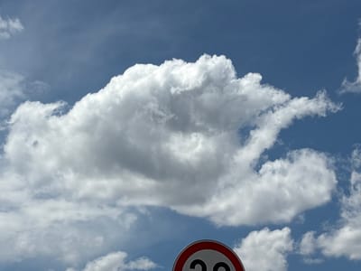 The image shows a clear blue sky with large, white, fluffy clouds. In the foreground, a circular traffic sign with a red border is visible at the bottom center of the image. The sign has the number "20" printed in black on a white background, indicating a speed limit of 20 kilometers per hour. The sign is partially obscured by clouds. The scene appears to be an outdoor environment, likely a road, given the presence of the speed limit sign. The location is identified as Yoakim Gruevo, Bulgaria.