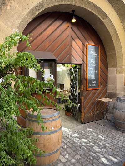 The image shows a wide, arched wooden doorway with a wrought-iron gate ajar, leading into a courtyard. The doorway is set within a stone archway, likely an entrance to a building. To the left of the doorway, a large wooden barrel stands prominently, partially obscured by lush green foliage with small white flowers. A second, similar barrel is visible to the right of the doorway.

A chalkboard sign is affixed to the wooden door, displaying opening hours in white chalk lettering. It reads:

"Unsere Öffnungs- zeiten

Dienstag - Freitag
16 - 23 Uhr

Samstag
11.30 - 23 Uhr

durchgängig warme
Küche
bis 21 Uhr"

In the foreground, the ground is paved with cobblestones, with patches of sunlight and shadow creating a dappled effect. A small, woven stool is placed near the right barrel. Inside the courtyard, visible through the open gate, are tables and chairs, suggesting an outdoor dining area for a restaurant or cafe. The architectural style and the presence of the barrels hint at a location with historical significance, possibly a wine cellar or a traditional establishment.

This scene is located at the "Bürgerspital Weingut" in Würzburg, Germany. The vineyard and restaurant are situated within the historic walls of the Würzburg city hospital.
