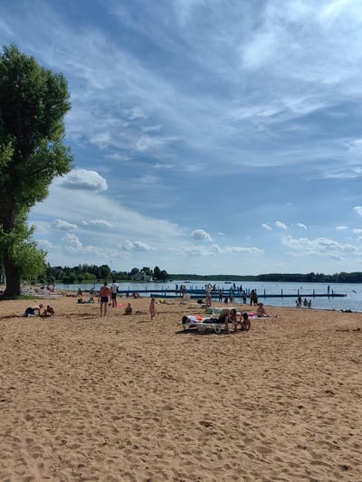An aerial view captures a sun-drenched beach scene at Lake Myadel in Myadzyel, Belarus. The sandy shore is populated with numerous people enjoying the summer weather. In the foreground, individuals are lounging on beach chairs or playing in the sand. Some are lying down, while others are interacting with each other. Further down the beach and closer to the water's edge, more people are visible, some wading into the lake or standing near a floating platform and a wooden pier. The lake itself appears calm with a few distant boats and people swimming. The background is characterized by a treeline and buildings, suggesting a developed area adjacent to the natural landscape. The sky is a vibrant blue, dotted with scattered white clouds. A large tree stands prominently on the left side of the image, framing the beach and water.