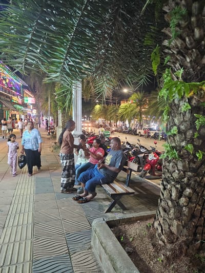 The image depicts a busy street scene at night in Ban Klong Haeng, Thailand. The foreground is dominated by a stone wall and lush green palm fronds. A group of people are seated on a wooden bench, with a man in a gray t-shirt and blue jeans in the center, looking down. Beside him, a woman in a pink shirt and a person in a patterned skirt are also seated. Further down the sidewalk, more people are walking and interacting.

The street is lined with shops and stalls, illuminated by colorful lights and signs, suggesting a commercial or entertainment district. Several motorcycles are parked along the roadside, indicative of local transportation. The overall atmosphere appears lively and active, typical of a nighttime scene in a tourist or local gathering area.

The lighting suggests it is evening or night. The presence of palm trees and the style of the buildings might suggest a tropical location, which aligns with the provided location context. There are no obvious weather cues, but the scene is well-lit, indicating artificial light sources are in use. The emotions of the people are difficult to discern due to the quality of the image and the lighting conditions.

Visible text includes what appears to be shop signs in Thai script, though the text is mostly illegible due to the image's resolution and angle. One sign in the distance reads "GOOD FOOD" in English.