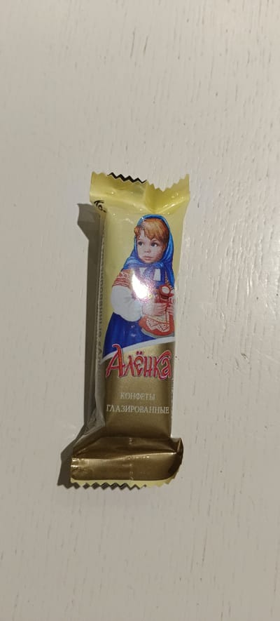 The image shows a single candy wrapper with a depiction of a young girl wearing a blue headscarf and a traditional Russian costume. The text on the wrapper is in Russian and reads "Алёнка" (Alyonka) and "КОНФЕТЫ ГЛАЗИРОВАННЫЕ" (Glazed Candies). The wrapper is a light yellow color with gold accents at the bottom. It is placed on a white, textured surface, likely a wooden table, and casts a shadow to the left. The lighting suggests an indoor setting. There is no discernible information about its location in Падлабенскі сельскі Савет, Belarus.