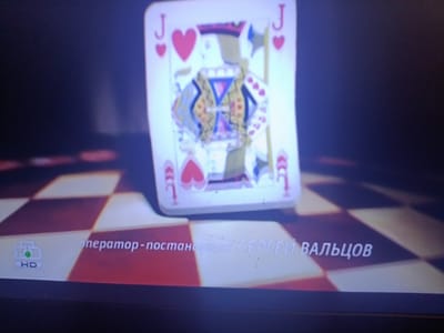 The image shows a close-up of a playing card, specifically the Jack of Hearts, on a blurred background. The card is upright and centered. Below the card, a checkered pattern is visible, resembling a chessboard. In the lower left corner, a logo with the letters "НТВ HD" is present. To the right of the logo, white text is displayed, which appears to be a caption or credit. The text reads "оператор-постановщик СЕРГЕЙ ВАЛЬЦОВ". The overall scene is dimly lit, with a spotlight effect on the playing card. There are no people or any discernible elements of the city of Александрыйскі сельскі Савет, Belarus.