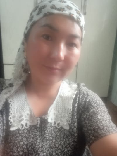 A medium shot captures a woman from the chest up, wearing a headscarf and a patterned dress with a lace collar. The woman's head is slightly tilted, and she is looking towards the camera. She has a neutral expression. The headscarf is white with a floral pattern. The dress is dark with a subtle floral print. The lace collar is white and has an intricate design. The background appears to be an indoor setting, with a neutral-colored wall. No specific location in Kerben, Kyrgyzstan can be identified from the image.