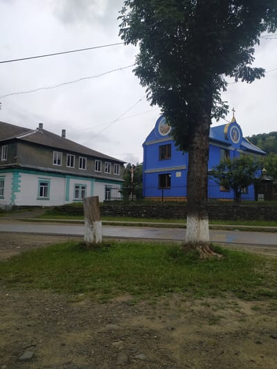 The image captures a scene in Bohdan, Ukraine, on a cloudy day. A large tree with a trunk painted white at its base stands in the foreground, to the right of center. Behind the tree, a bright blue building with a gold-accented roof and circular religious icons on its facade is visible. To the left of the blue building is a longer, two-story building with a grey roof and white and green lower walls. The buildings appear to be situated near a road, with a grassy area and a rough patch of ground between the road and the buildings. A wooden post, also painted white at its base, stands in the grassy area to the left. Further to the right, behind the blue building, a small wooden structure with a golden dome can be seen, and a hillside covered in trees rises in the background. A crucifix with a figure of Christ is visible near the fence of the longer building on the left. The overall impression is of a village or town setting, likely with religious significance.