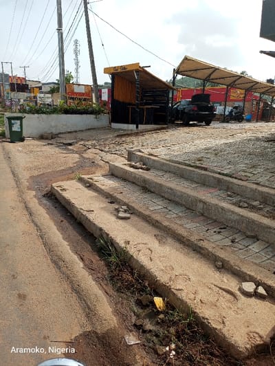 The image shows a set of concrete steps leading up to a paved area in Ado Ekiti, Nigeria. The steps are worn and cracked, and there is a ditch filled with dirt and debris to the left of the steps. A single green trash can is located near the edge of the ditch. The background shows a street lined with buildings, including a small structure with a sign that reads "Snack Station." A silver car and a motorcycle are parked in the paved area to the right of the steps. The street is lined with power lines. The sky is partly cloudy. The coordinates (latitude: 7.649, longitude: 5.226) indicate a location in the city of Ado Ekiti, Nigeria.