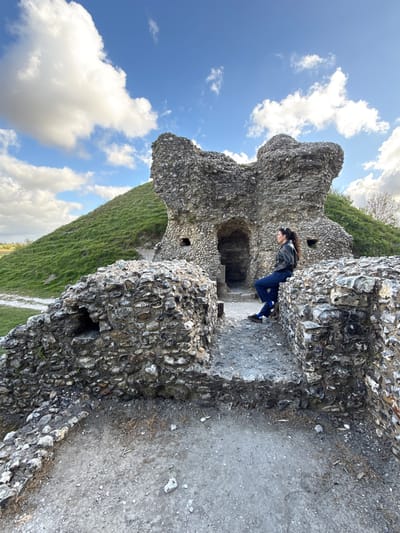 The image depicts a person exploring ancient ruins under a clear blue sky with scattered white clouds, likely situated in King's Lynn and West Norfolk, United Kingdom. The scene features weathered stone structures, possibly a castle or other historical building, constructed from rough, grey-brown stones, characteristic of flint masonry.
A young woman with long dark hair is the primary subject, standing within the ruins. She is dressed in a dark leather jacket, dark blue jeans, and dark shoes with white soles. Her posture suggests she is observing her surroundings or pausing during an exploration of the site, leaning slightly forward and holding onto a section of the stone wall. Behind her, a dark arched opening indicates a passage or chamber within the ruins.
The ground around the ruins is composed of earth and gravel. Beyond the immediate structures, a verdant green grassy hill rises, extending into the background. The bright daylight suggests it is daytime, possibly late morning or early afternoon, with fair weather conducive to outdoor activities. The image conveys a sense of historical exploration and tranquility. There is no visible text present in the media.