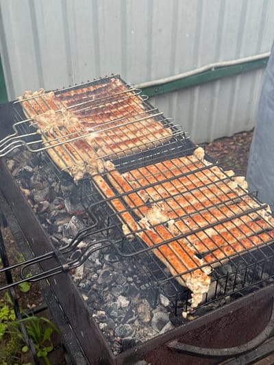 The image depicts a close-up, outdoor shot of sausages grilling on a charcoal barbecue. Two large grill baskets are filled with numerous sausages, some appearing browned and cooked, while others are lighter in color. Embers and ash are visible beneath the grill. A grey corrugated metal fence or wall forms the background, with some greenery and what appears to be a person's leg and gray fabric on the right side of the frame. The scene is consistent with a residential outdoor setting, possibly a dacha or backyard in Газета Труд Снт, Russia.