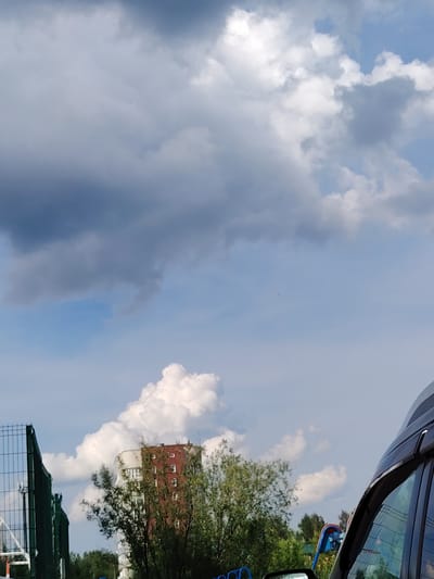 The image shows a street scene in Pyt'-Yakh, Russia, under a cloudy sky. In the foreground, a dark-colored vehicle is partially visible on the right side. To the left, a green sports fence is present. Behind the fence and vehicle, a multi-story building with red and white elements is seen, surrounded by trees. The sky is filled with a mix of dark gray and bright white clouds, with patches of blue sky visible.