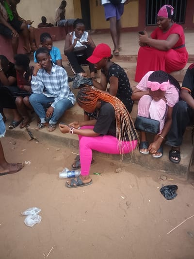 In Umuokpu, Nigeria, an outdoor scene depicts a group of approximately ten individuals, primarily young adults and women, gathered near a building with a light brown facade and concrete steps. The ground is composed of compacted dirt and sand, showing scattered litter including discarded plastic bottles and wrappers.
In the immediate foreground, a young woman with long, braided orange-brown hair, wearing a black t-shirt, bright pink leggings, and grey sandals, is crouched on the ground, actively engaged with a mobile phone held in her hands. A clear plastic water bottle lies on the ground near her feet.
On the concrete steps to the right, a woman in a red dress and a pink and black patterned headscarf is seated, also looking at a mobile phone. Below her, another woman in a light pink top and trousers is hunched over, looking downwards, with a black handbag positioned between her legs. Partially visible next to her is a person in dark trousers and black sandals.
On the steps towards the center-left, a young woman wearing a blue plaid shirt and blue jeans sits, looking forward with a neutral expression, a folded item resembling a magazine or book resting on her lap. To her right (viewer's left), another young woman in a black t-shirt and a red baseball cap is seated, looking down. Further left, a woman in a white patterned top is seated, focused on an object in her hands, likely a mobile phone.
In the background, several other individuals, including men, are seated on steps or partially visible standing. Most individuals are wearing sandals or slippers. The general posture of the group suggests waiting or resting. No direct interactions between individuals are discernible, with many focused on personal devices or their immediate surroundings.