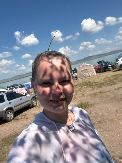 A woman is captured in a selfie against a backdrop of a bright blue sky with scattered white clouds, a body of water, and a shoreline dotted with tents and vehicles. The woman, with her hair pulled back, wears a sheer white top that allows the dappled sunlight to cast shadows across her skin. A small plant sprout is attached to a hair clip, emerging from the top of her head like an antenna. In the background, several cars and tents are visible, suggesting an outdoor gathering or campsite. The environment appears to be a natural landscape with a body of water and distant hills under a clear, sunny sky. The specific location is the city of Zhemchuzhny, Russia.