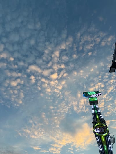 The image shows a tall amusement park ride with flashing lights against a sky filled with scattered clouds. The ride is green and black with circular and rectangular sections illuminated by bright white, green, and blue LEDs. The number "360" is prominently displayed on a circular section of the ride, also lit by LEDs. The sky transitions from a pale blue at the top to warmer, yellowish tones near the bottom, suggesting either sunrise or sunset. In the upper right corner, a black fixture, likely a light or part of another ride, is visible. The scene appears to be set at an outdoor amusement park, possibly in Golubitskaya, Russia, given the context provided. There are no people visible in the image.
