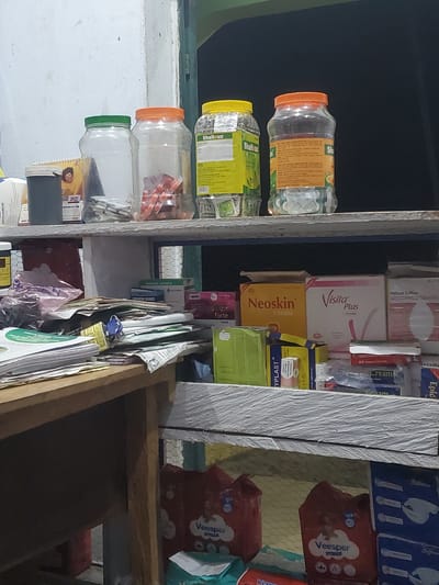 This is a medium shot, eye-level photograph taken in a retail setting, likely a pharmacy or small shop, in Mutum Biyu, Nigeria. The image depicts a cluttered shelf filled with various pharmaceutical products and supplies.
In the background, three large, clear plastic jars with colorful lids (green, orange, and yellow) contain what appear to be pills or capsules. Labels on these jars indicate brands like "Shaltlux." To the left of the jars, a smaller dark grey container sits next to a spiral-bound calendar showing the year 2025 with a picture of a smiling person.
The middle shelf is crowded with stacked items. A collection of papers and documents is piled on the left side, some held together by a blue lock and key. Various boxes of medical supplies are visible, including "Neoskin Cream," "Visita Plus Cream," and "Fortune Saffron Ketoconazole Cream BP." There are also smaller boxes labeled "TYPLAST" and others containing fabric plasters and surgical blades.
The lower shelf displays larger packages of what appear to be sanitary products or diapers, branded "Veesper." Boxes of other items, possibly pharmaceutical or hygiene products, are stacked to the right. The shelves themselves appear to be made of wood and possibly metal, with a textured white surface. The overall environment suggests a busy, functional space where goods are stored and displayed for sale. The lighting is somewhat dim, contributing to a slightly shadowy atmosphere.