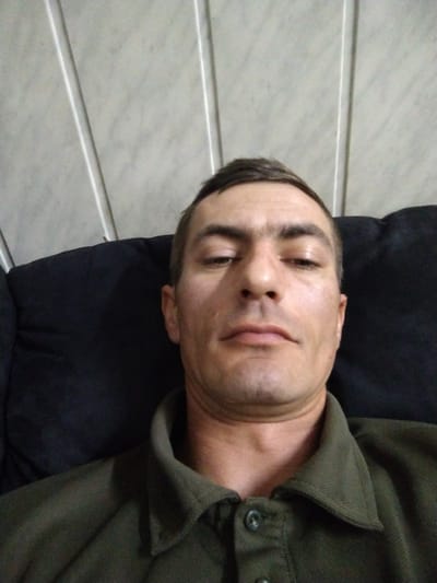 A man, appearing to be in his late 30s or early 40s, is captured in a close-up, slightly upward-angled selfie. He has short, dark brown hair, fair skin with some blemishes, dark eyebrows, and is looking directly at the camera with a neutral expression. He is wearing a dark green collared shirt, and the background consists of a black upholstered surface, likely a couch or chair, and a light-colored wall with vertical paneling. The image was taken indoors. No specific landmarks or features in the image definitively place the location in Vylok, Ukraine.