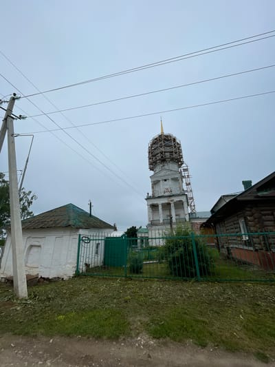The image shows a view of a church bell tower under scaffolding, with surrounding buildings and infrastructure in Кстинино, Russia. A white, multi-story bell tower with classical architectural elements, including columns and arched windows, stands prominently in the center-right of the frame. The upper portion of the tower is covered in a wooden scaffolding structure, suggesting ongoing construction or restoration. A golden spire tops the bell tower. To the left of the bell tower is a small, white, single-story building with a green, weathered, sloped roof. A green metal fence encloses the area in front of this building and extends towards the bell tower. To the right of the bell tower, a portion of a dark, wooden log house is visible. Overhead, power lines crisscross the cloudy, gray sky, supported by a concrete utility pole on the left side of the image. The ground in the foreground is unpaved, with a dirt and gravel surface transitioning into a strip of green grass.