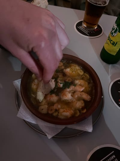 A hand is dipping a piece of bread into a bowl of shrimp and garlic in a clay bowl, sitting on a white napkin on a gray table. The bowl sits on a gray plate. The hand is out of focus, and the table is also out of focus, but the bowl and the food are in focus. The bowl is full of shrimp and garlic, and there is some parsley sprinkled on top. There is a glass of beer and a bottle of beer in the background, out of focus. The beer bottle is green, and the beer is amber. The location is in a restaurant in Arucas, Spain, based on coordinates 28.118 latitude, -15.524 longitude.