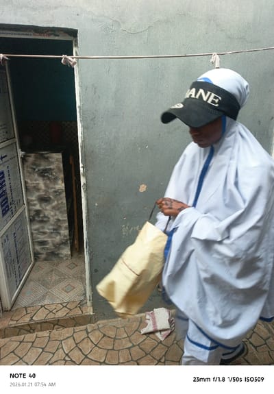A woman wearing a white robe with blue trim and a black baseball cap is walking. She is carrying a light yellow bag. The backdrop includes a weathered building with an open doorway revealing an interior. The steps leading up to the doorway have patterned tiles. A clothesline is strung across the scene. The image details indicate it was taken with a 23mm lens at f/1.8, 1/50s shutter speed, and ISO 509. The date is marked as January 21, 2026, at 07:54 AM.