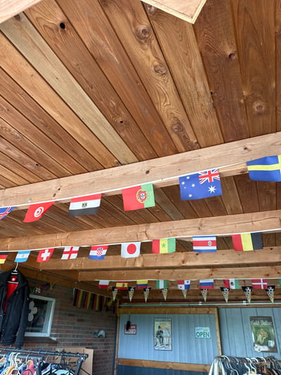 This image captures a static scene within an outdoor or semi-sheltered area in Deinste, Germany, likely a retail space such as a market stall or second-hand shop. The overhead structure consists of a wooden plank ceiling featuring distinct wood grain and knots, with a prominent horizontal wooden beam running across the middle.

Numerous strings of international flags are suspended from the wooden beams. The top row, from left to right, displays flags of the United Kingdom (Union Jack), Tunisia, Egypt, Portugal, Australia, and a partially visible flag of Sweden. The middle row, from left to right, includes partially visible Germany, Argentina, Switzerland, England, Croatia, Japan, Senegal, Costa Rica, and partially visible Belgium. Below these, a third string features small, inverted cone-shaped pennants with soccer ball designs interspersed with flags of Colombia, Nigeria, Panama, Russia, Mexico, and Denmark.

To the left, a dark jacket with a red interior and a white logo (resembling a Swiss cross) on the chest is hanging on a hanger. Below it, a small, white-framed rectangular window is set into a red-brown brick wall, with a white, decorative cutout (possibly a flower) visible on the interior side. A white object, possibly a hat or helmet, is hanging on the brick wall to the right of the window.

The lower wall section on the right consists of light blue-grey vertical paneling or corrugated material. A matching light blue-grey wooden door, possibly a stable door, is adorned with two signs: a red-framed "WE'RE CLOSED" sign and a green-framed "OPEN" sign. Two vintage-style posters are affixed to the blue-grey wall: one on the left for "LESONJE-CAKES" depicting a seated man, and one on the right for "CIGARETTES Gold Dollar" featuring a woman and the text "20 pieces."

In the foreground, two metal clothing racks are visible, laden with various garments on hangers, suggesting a display or sales area. No individuals are present in the scene, and no specific actions or interactions are depicted.