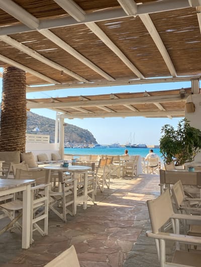The image depicts an outdoor dining area of a restaurant or cafe in Φάρος, Greece, overlooking a sunlit bay. The scene is brightly lit under a clear blue sky, with the turquoise water of the bay visible in the mid-ground. Rocky hills form the coastline in the background, with a small white building prominent on the left hillside.
The foreground is dominated by the restaurant's structure, which includes a white wooden pergola-style roof covered with natural reed or bamboo thatch. A large, textured column, appearing to be wrapped in palm fiber, is on the far left. The flooring consists of irregular reddish-brown flagstones, marked by distinct shadows from the overhead structure.
The dining area is furnished with numerous white wooden tables, both round and rectangular, and matching white wooden chairs, some featuring light-colored fabric seating. On the far left, a white built-in bench is adorned with light-colored cushions and pillows. A potted plant with green foliage and light-colored fruit rests in a white pot on the right side. Small blue containers or cups are visible on several tables. Many tables and chairs in the foreground are unoccupied.
Two individuals are visible in the mid-ground, seated at a white table facing the water. The person on the left is seen from behind, with dark hair. The person on the right is also seen from behind, wearing a light-colored short-sleeved shirt with the word "CLUB" partially visible on the back. Both appear to be observing the marine activity.
The bay is populated with various vessels: multiple large white yachts are anchored or moored, numerous smaller boats are docked in a marina to the left, a large white catamaran-style ferry is visible further out, and the masts of several sailboats pierce the horizon.