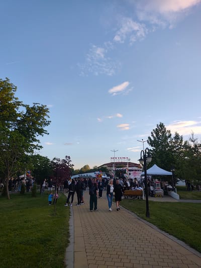 The image depicts a public event in Bozhurishte, Bulgaria, near the "SPETEMA HOTEL". A paved path leads through a grassy area with trees on either side. Many people are present, with a crowd gathering in the distance near a stage or event area decorated with flags and balloons. A white tent is set up on the right side of the path. A pair of people walk toward the viewer on the path. A small child stands by a tree on the left. The sky is blue with scattered clouds. A contrail can be seen in the upper left.