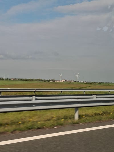 The image shows a view from a vehicle moving on a highway. A gray guardrail is visible on the right. To the right of the guardrail is a grassy area with some yellow flowers, adjacent to the highway's asphalt road surface with a white lane marker. The left side of the frame captures a cloudy sky, transitioning into a landscape with wind turbines in the distance. Based on the prompt the location is Palauzovo, Bulgaria.