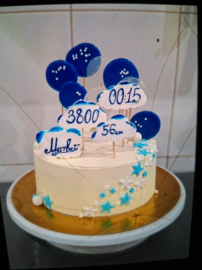 A frosted cake decorated with blue and white cloud-shaped toppers, round blue balloons, and star-shaped sprinkles is displayed on a gold cake board. The cloud toppers feature writing and numbers, including "Матвей" (Matvey), "3800", "56см" (56 cm), and "00:15". The cake itself is decorated with blue and white stars. The background appears to be a wall with a grid pattern, possibly tiled. The image is slightly distorted and has visible cracks, suggesting it may be a photograph of a screen or a damaged print. The location of Medvezhyi Ozyora, Russia is not evident in the visual information provided.
