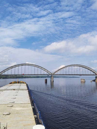 The image depicts a large, flat-topped barge traveling on a wide body of water, possibly a river or lake, under a double-arched bridge. The bridge is constructed of steel arches supported by concrete pillars and suspension cables. There are remnants of old pillars of the bridge on the river. The barge has a blue railing and a surface with numerous circular access points or hatches. Behind the bridge, a strip of forested land is visible, separating the water from the horizon. The sky is blue with numerous scattered white clouds. The water is dark and rippled. The scene's location cannot be conclusively confirmed as Zarya, Russia based on the provided image alone.