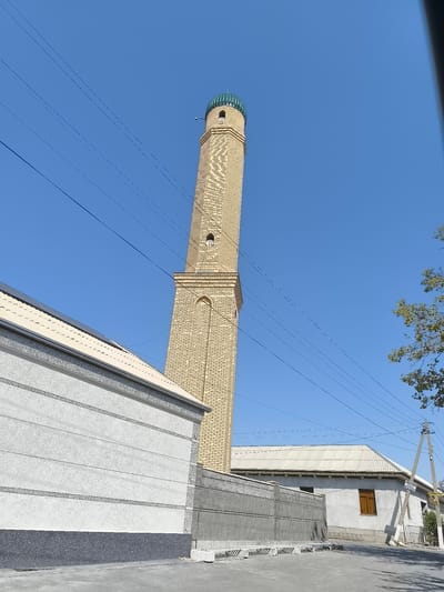 A minaret with a turquoise dome stands tall against a clear blue sky in Babakhodja, Uzbekistan. The minaret is constructed of light yellow bricks and features decorative brickwork and arched openings. In the foreground, a white building with a gray textured lower section is visible, with a sloping roof that appears to have solar panels. A gray concrete fence separates this building from a street, where utility poles and overhead power lines are present. To the right of the minaret, another building with a light-colored, corrugated roof is visible, along with a single brown window and a fence. A tree with green foliage is also present on the right side of the image. The overall scene depicts a street view in a town or village setting.