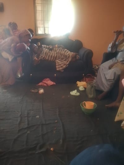 The image depicts an indoor scene in the city of Fanda, Nigeria, likely a residential living area. The room features light orange walls and a window with horizontal and vertical bars on the upper left, emitting bright natural light that causes a significant glare. The floor is covered with a dark fabric sheet or mat, showing scattered debris.

Multiple individuals are present. On the far left, two figures are partially visible, one appearing to hold another person. In the center-left, an individual wearing a light-colored, striped garment lies on their left side on a dark sofa, holding and operating a white smartphone. To the right, another individual, dressed in a light-colored flowing garment, sits on a sofa, holding a dark cylindrical object near their mouth. The lower right foreground shows the leg and foot of another person, indicating their presence on the floor.

On the floor, various items are scattered, including a red patterned fabric, a small light-colored object, a pair of light green open-toed sandals, a light-colored handbag, and a green bowl containing an orange mug.