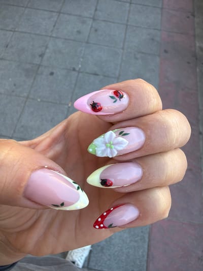 A manicured hand is presented against an outdoor background of a grey and pink brick walkway. The fingernails are almond shaped and painted with a glossy light pink base. The nail on the index finger features two red ladybugs with black spots and green leaves. The middle fingernail has a 3D white flower with light green accents. The ring fingernail has a yellow tip with a single ladybug. The pinky fingernail has a red tip with white polka dots. The thumb has a white tip with small green leaf designs. The skin of the hand appears lightly tanned with visible wrinkles and pores. The location appears to be in Azuqueca De Henares, Spain based on the location data associated with this image.