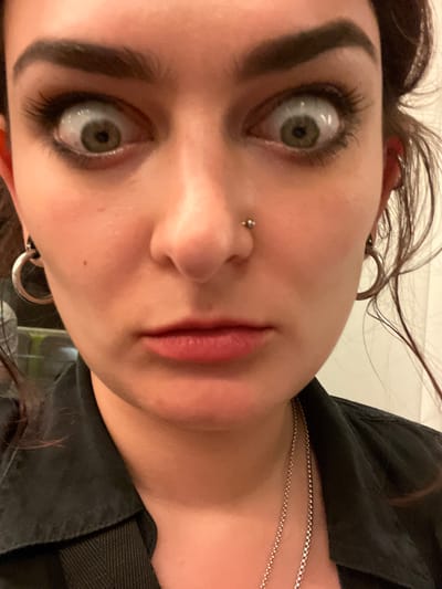 A close-up, slightly tilted, high-angle photograph depicts a person with fair skin. The individual has prominent, dark eyebrows, long eyelashes, and light-colored eyes that appear to be gazing upwards. A small stud is visible in the left nostril. The lips are visible and appear to have a neutral expression. Parts of hoop earrings are visible in the ears. Stray strands of hair are visible around the face. A silver necklace rests on the person's collarbone, and a portion of what appears to be a black jacket is also visible. The background is plain and suggests a neutral indoor environment. There are no indications of the image being taken in Torrent, Spain.