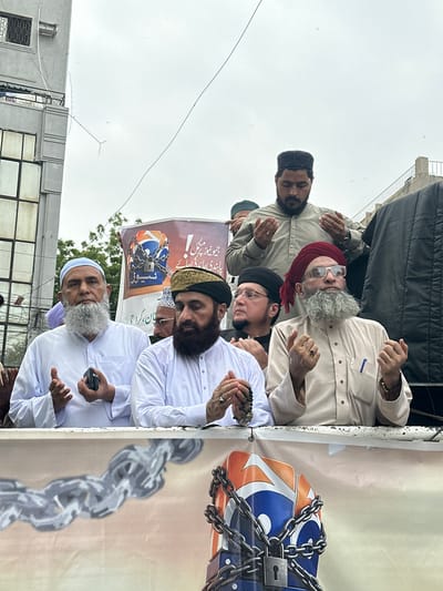 This media file captures a group of men gathered outdoors, possibly for a religious or political event. The subjects are primarily men, many of whom are wearing traditional Islamic attire, including caps and long robes. Some have beards, and one individual wears a red turban and glasses. The setting appears to be urban, with buildings and a clear sky visible in the background, suggesting it is daytime. The weather is overcast, indicated by the gray, diffused light.
The men are shown in various poses, with several having their hands raised in prayer or supplication. Some are looking upwards, while others are engaged with each other. One man is holding a mobile phone. There is a large banner or backdrop behind the group, featuring graphics of chains and a locked object, possibly symbolizing oppression or imprisonment. The banner also contains text in what appears to be Urdu. The overall mood seems serious and possibly somber, given the expressions and the imagery on the banner. The location is identified as Karachi Division, Pakistan.